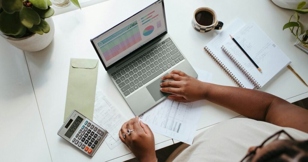 Gestora analisa planilha de orçamento no notebook com calculadora, recibos e caderno de anotações sobre a mesa
