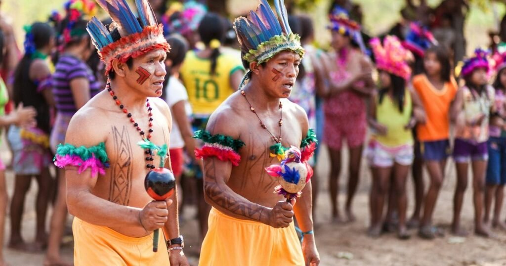 Homens indígenas com cocar de penas conduzem celebração tradicional na aldeia, com crianças e jovens da comunidade ao fundo