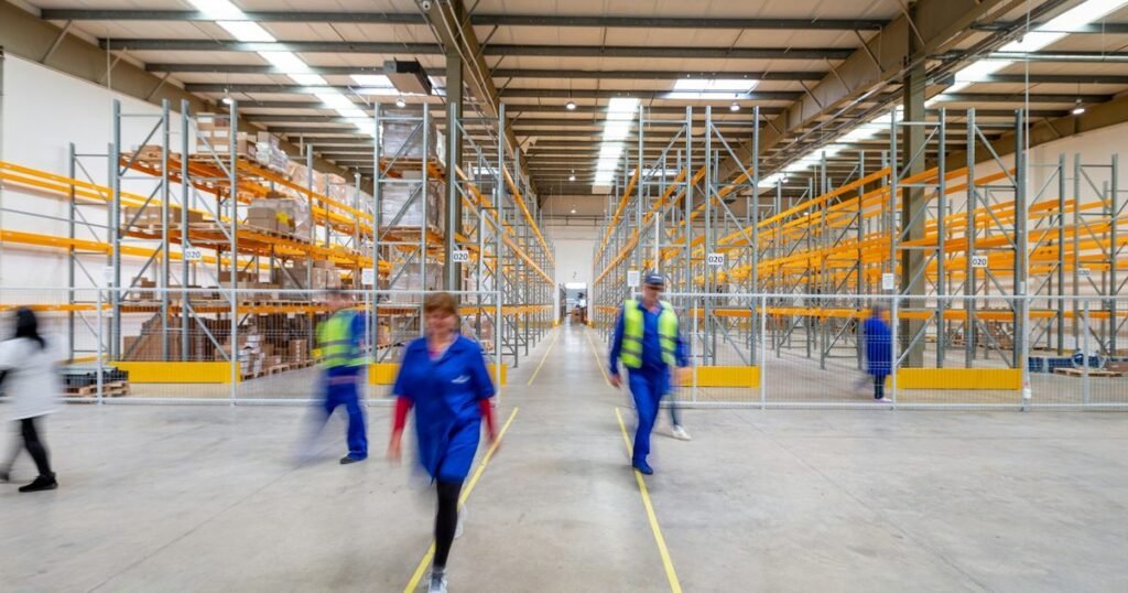 Profissionais de uniforme azul e colete caminham entre as estantes altas de um centro de distribuição moderno e iluminado