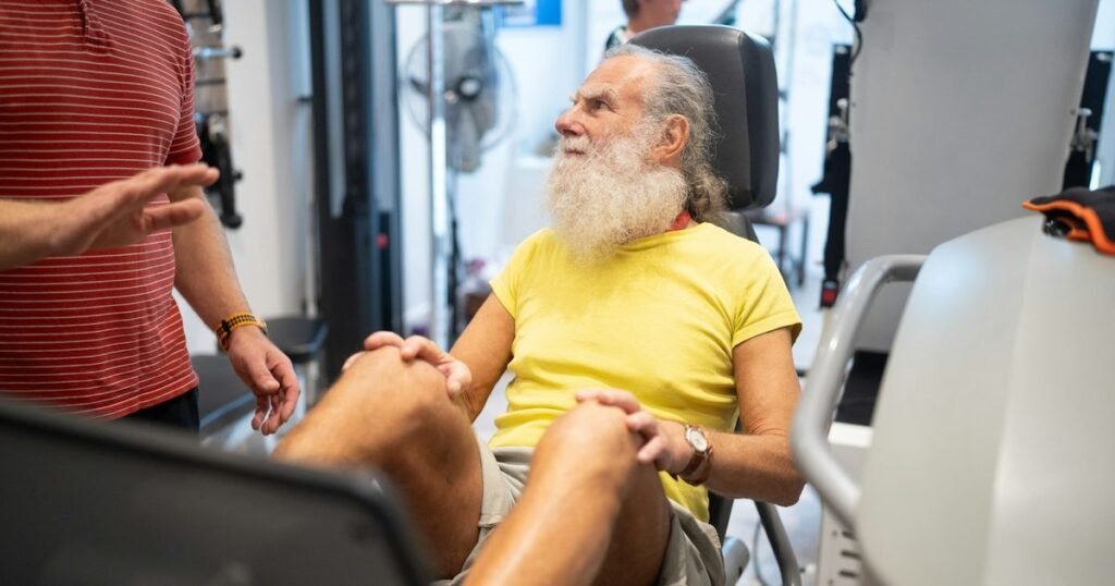 Idoso de barba branca em equipamento de reabilitação ouve a orientação do fisioterapeuta na academia terapêutica