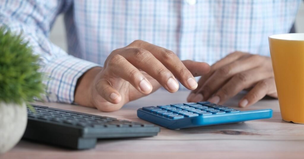 Contador digita em calculadora azul ao lado de teclado e caneca amarela sobre a mesa de trabalho