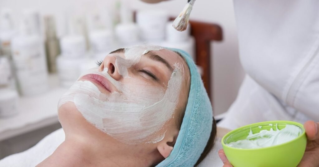 Esteticista aplica máscara facial com pincel no rosto de cliente deitada com touca de toalha, em clínica de estética com prateleira de cosméticos ao fundo