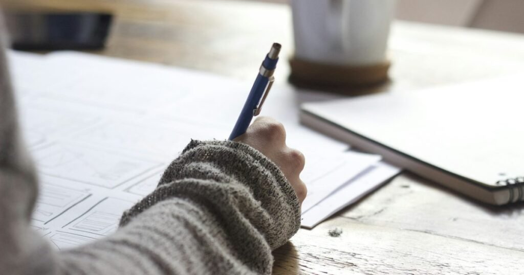 Mão escreve com caneta azul em folhas de papel sobre mesa de madeira, ao lado de caderno e caneca