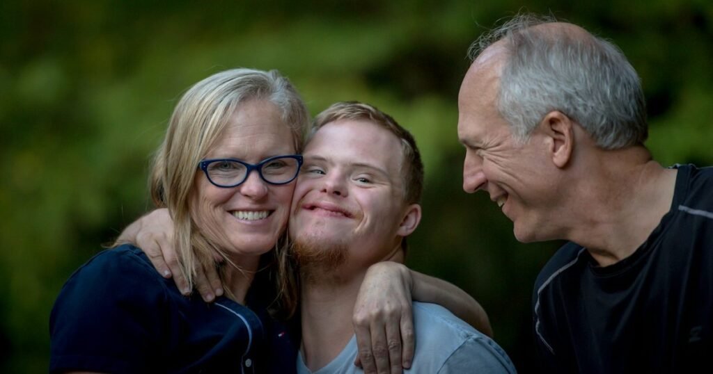 Pai e mãe abraçam o filho jovem com síndrome de Down, os três sorrindo, em área verde ao ar livre