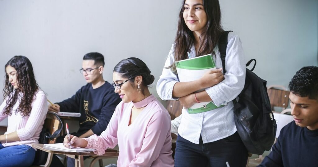 Sala de aula clara com jovens estudantes fazendo anotações nas carteiras e uma aluna em pé segurando um caderno verde