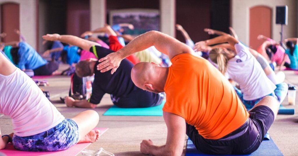 Aula coletiva de alongamento: pessoas de várias idades se exercitam sentadas em colchonetes coloridos, com um homem de camiseta laranja em primeiro plano