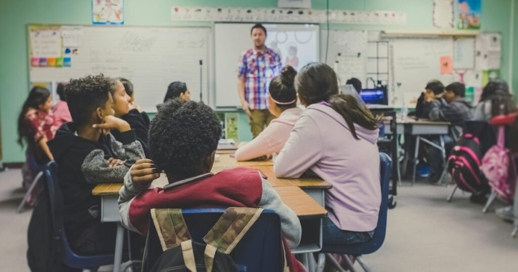 Professor em sala de aula conduzindo os alunos, com mochilas apoiadas nas carteiras, representando a gestão escolar no Brasil